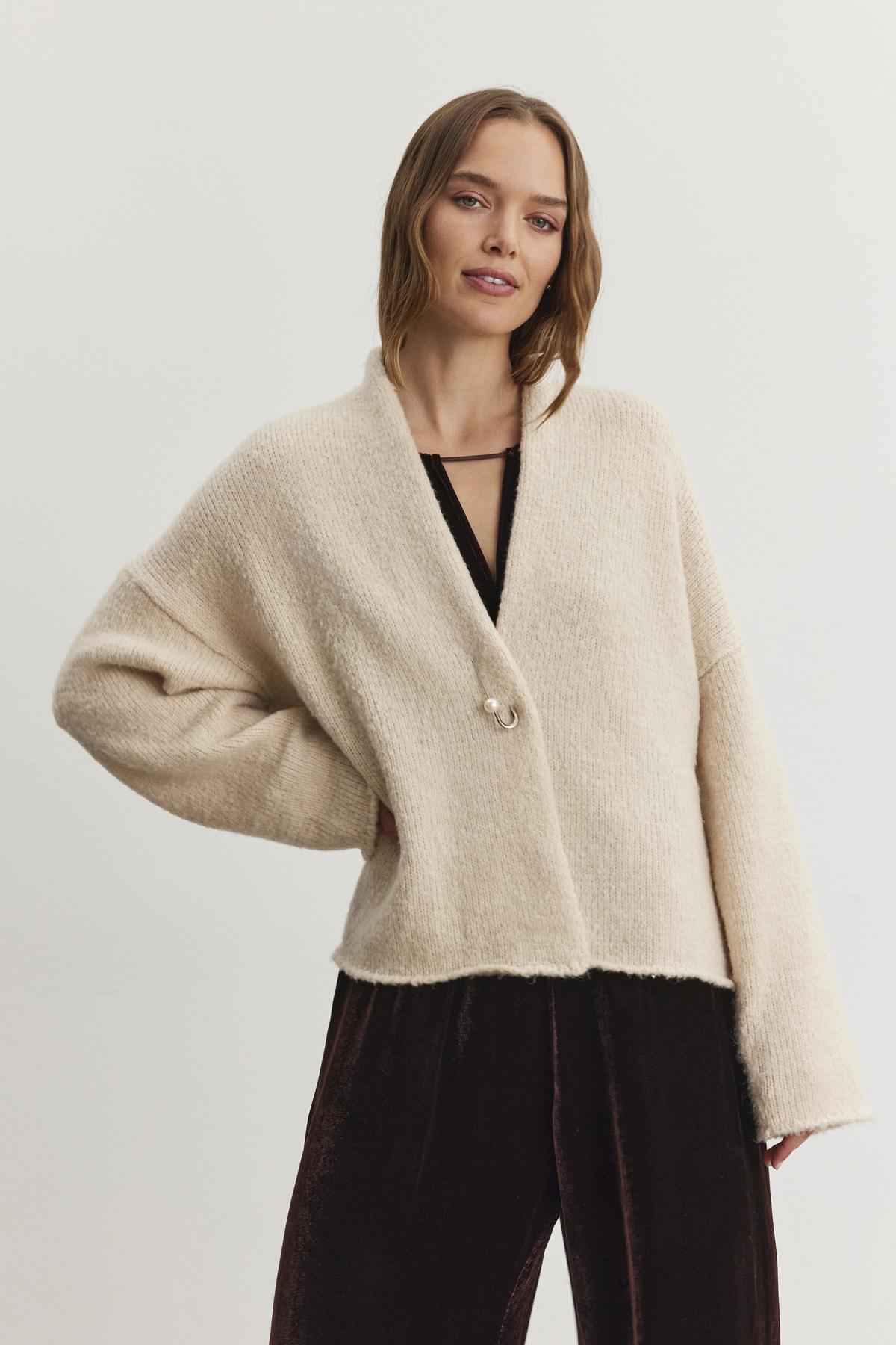   A person adorned in the CHANSON CARDIGAN by Velvet by Graham & Spencer, featuring a cream-colored and open-front design, stands against a plain white background over a black outfit. 