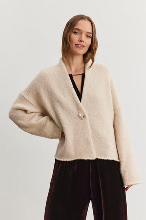 A person adorned in the CHANSON CARDIGAN by Velvet by Graham & Spencer, featuring a cream-colored and open-front design, stands against a plain white background over a black outfit.