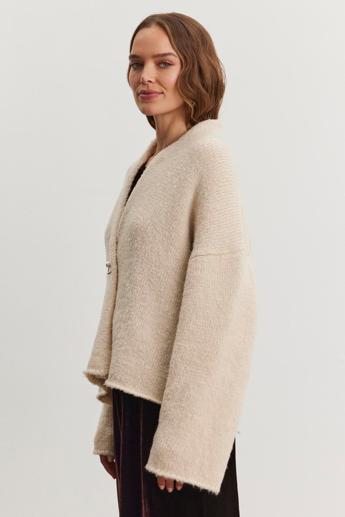   A person with shoulder-length hair is wearing the CHANSON CARDIGAN by Velvet by Graham & Spencer—a light beige, open-front cardigan featuring a cozy shawl collar—over a dark dress, standing against a plain background to highlight the added elegance of the ensemble. 