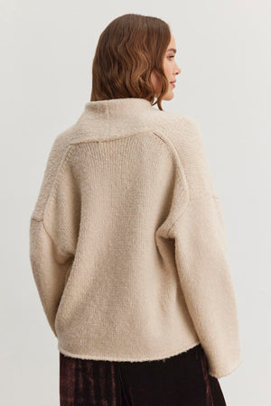 The individual is enveloped in the CHANSON CARDIGAN, an oversized beige knitted sweater featuring a shawl collar, by Velvet by Graham & Spencer, facing away.