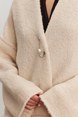 A person wearing the CHANSON CARDIGAN by Velvet by Graham & Spencer, a beige wool blend sweater with a pearl hook clasp and a shawl collar.