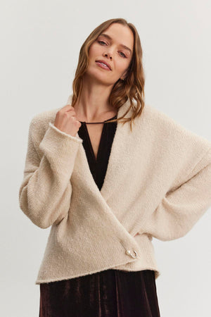 A woman wears the CHANSON CARDIGAN by Velvet by Graham & Spencer, a beige wrap sweater with a shawl collar, layered over a black dress as she poses against a plain white backdrop.