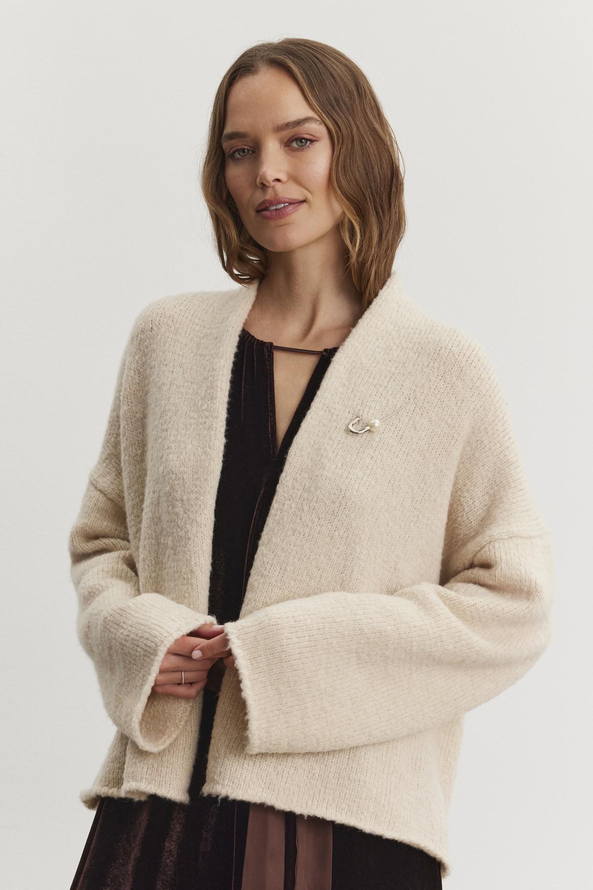 A person wearing the CHANSON CARDIGAN by Velvet by Graham & Spencer—a cream-colored, open-front cardigan with a shawl collar—over a dark dress stands in front of a plain background, looking at the camera.-38246388564161