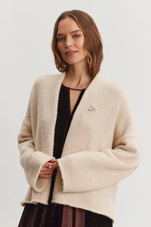 A person wearing the CHANSON CARDIGAN by Velvet by Graham & Spencer—a cream-colored, open-front cardigan with a shawl collar—over a dark dress stands in front of a plain background, looking at the camera.