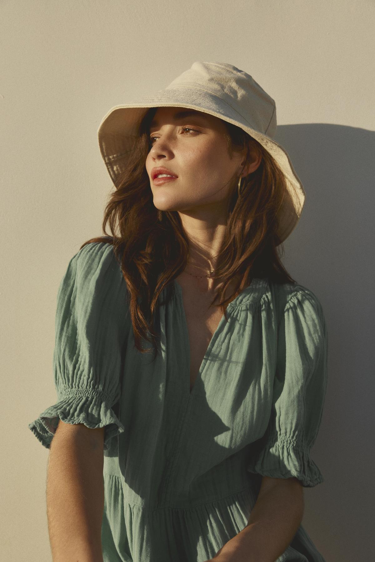 A woman with long brown hair is wearing a light green blouse and the Hazel Canvas Bucket Hat by Freya, made from sustainable craftsmanship, looking to the side while standing against a light-colored wall.-36443559755969