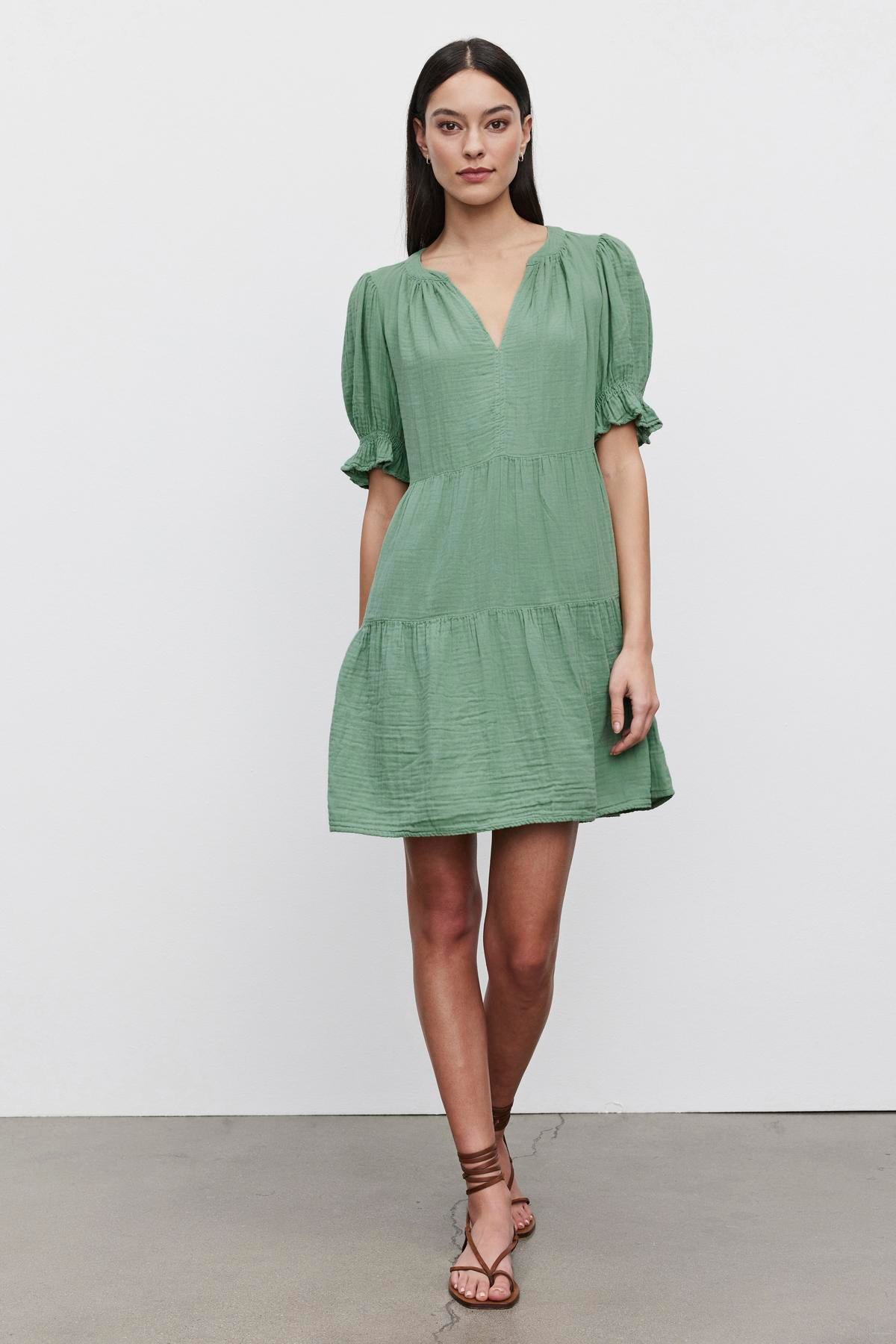 A woman stands against a plain backdrop, wearing a green, knee-length CLARISSA COTTON GAUZE TIERED DRESS with puffed sleeves and brown strappy sandals by Velvet by Graham & Spencer.-36426050699457