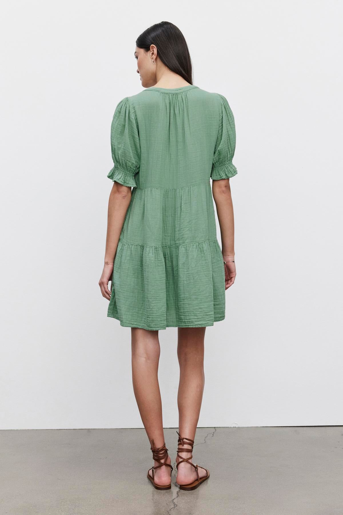   A woman in a green, short-sleeved, Velvet by Graham & Spencer CLARISSA COTTON GAUZE TIERED DRESS stands with her back to the camera, shown against a plain wall, wearing brown strappy sandals. 