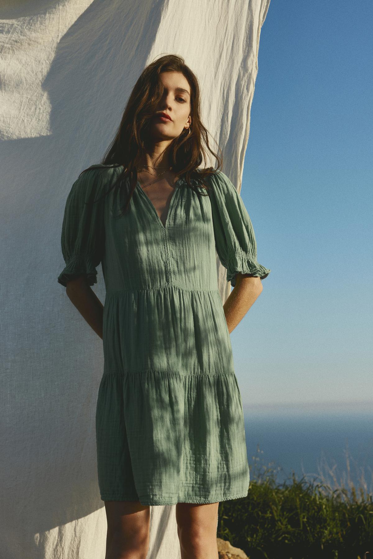   A woman in a Velvet by Graham & Spencer CLARISSA COTTON GAUZE TIERED DRESS with puffed sleeves stands beside a flowing white fabric with a sea background under clear skies. 