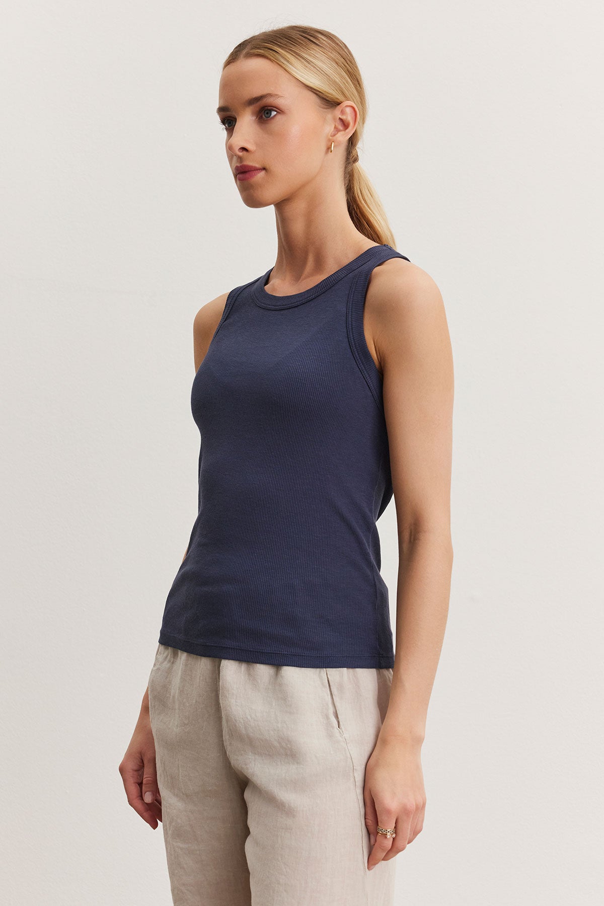   A woman wearing a fitted Velvet by Graham & Spencer NOLA tank top and beige trousers stands against a light background, looking to the side. 