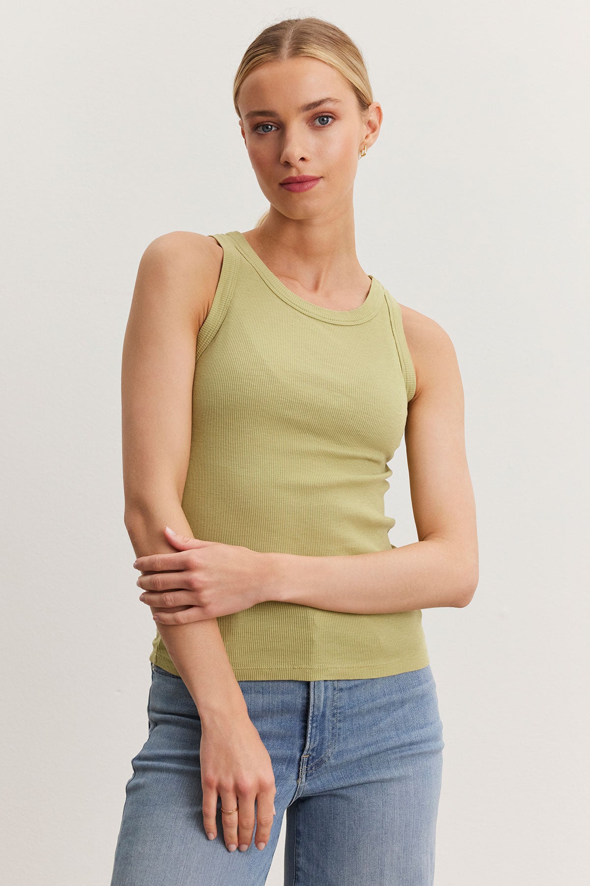   A woman in a Velvet by Graham & Spencer NOLA tank top and blue jeans stands against a plain background, posing with one hand on her hip and the other on her arm. 