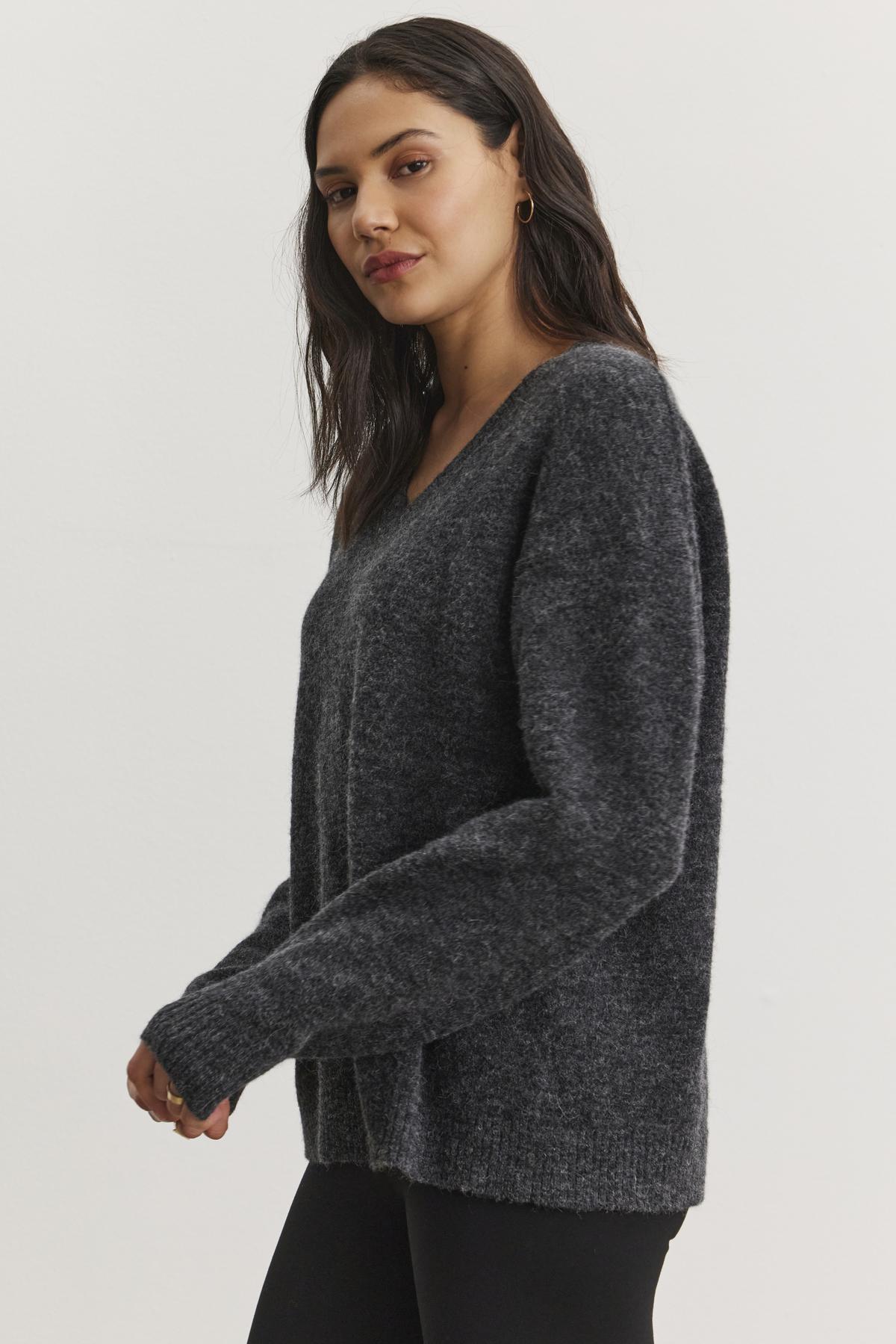   A person wearing the KELSA ALPACA SWEATER in dark gray from Velvet by Graham & Spencer and black pants stands against a plain white background, looking towards the camera. 