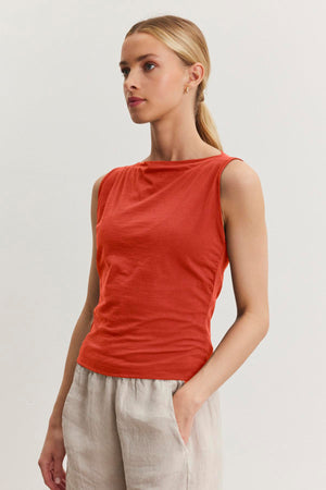 A person with blonde hair tied back is wearing a sleeveless, red EMILIA TANK TOP made of soft cotton and beige pants, standing against a plain, white background—perfect for casual wear with a modern minimal design by Velvet by Graham & Spencer.