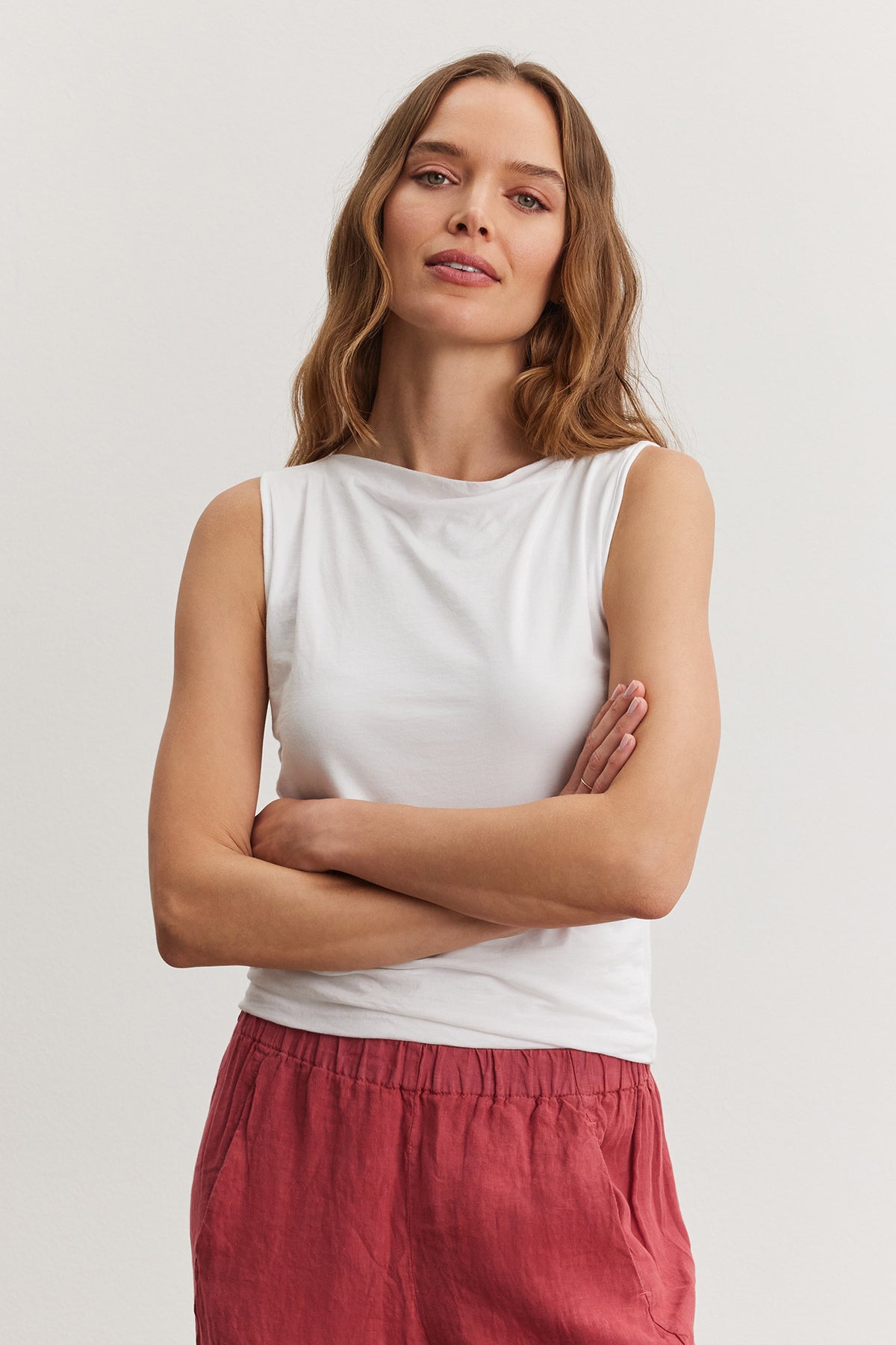 A woman with medium-length hair, wearing a Velvet by Graham & Spencer EMILIA TANK TOP and red pants, stands with her arms crossed.-36909542768833