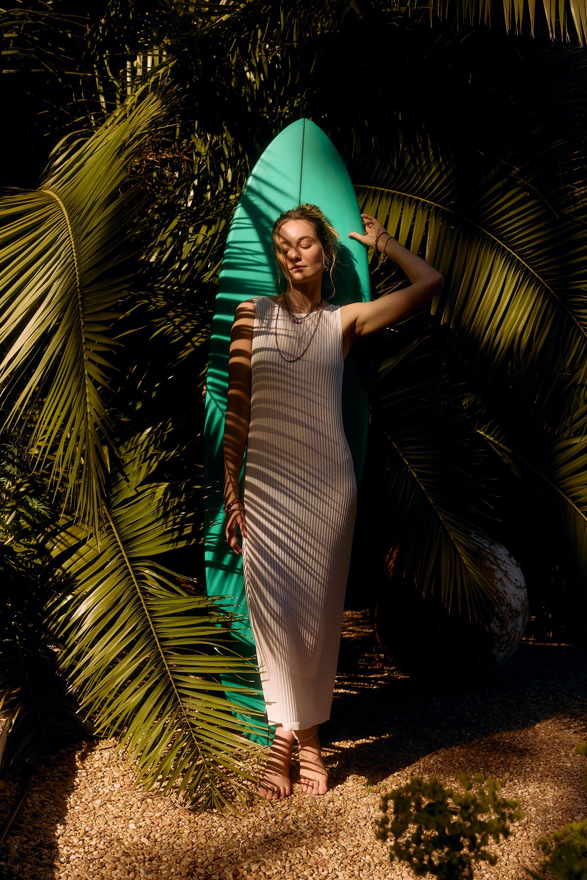 A woman in a white CALLEN DRESS leans against a turquoise surfboard surrounded by tropical plants by Velvet by Graham & Spencer.-36909770342593