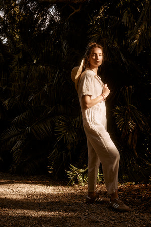 A woman in a casual white Velvet by Graham & Spencer Delilah linen jumpsuit and a sun hat stands among shadowy trees, sunlight highlighting her face and body.