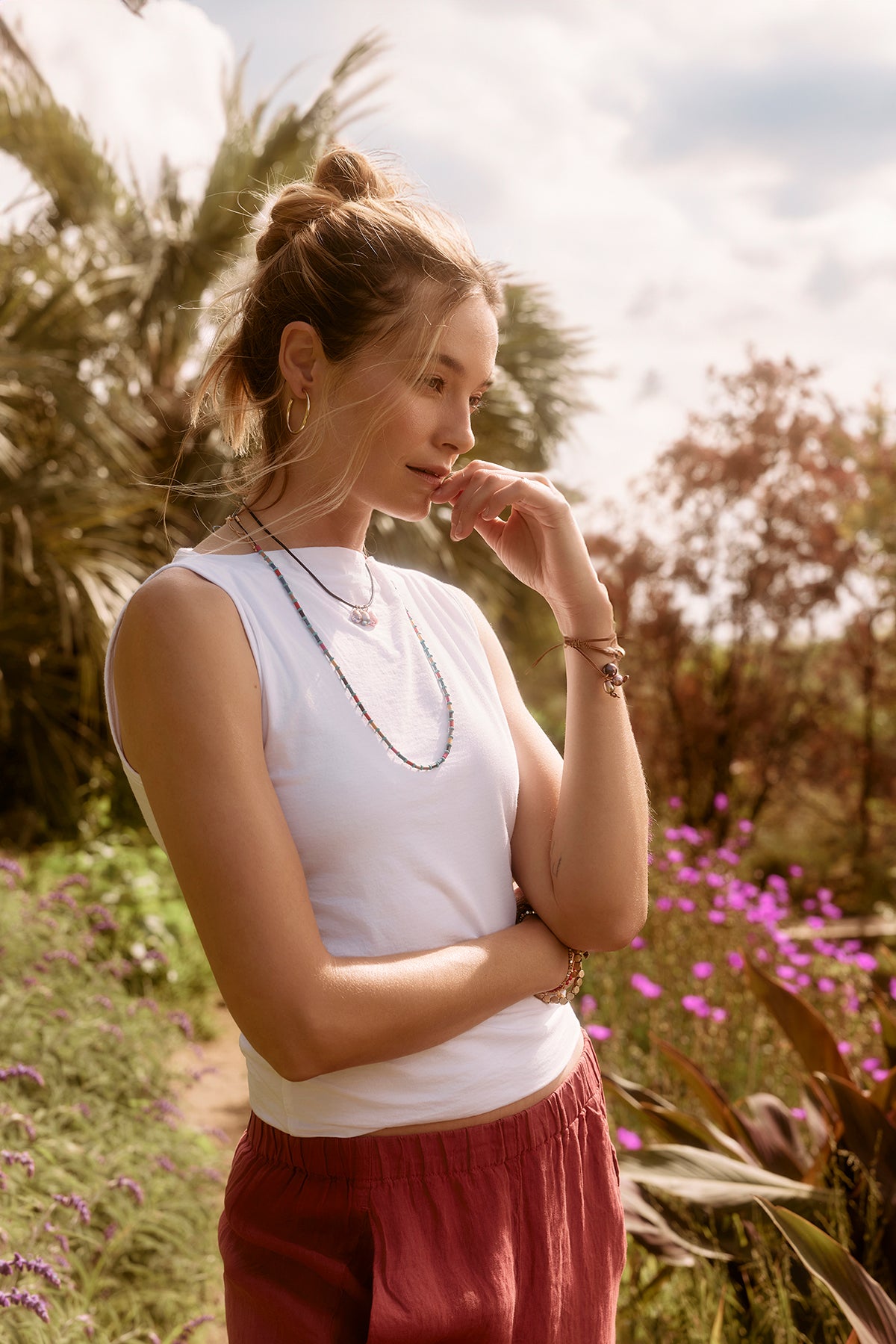 A woman in a Velvet by Graham & Spencer EMILIA TANK TOP and red pants stands pensively in a sunny field with purple flowers, her hand resting on her chin.-36909778403521