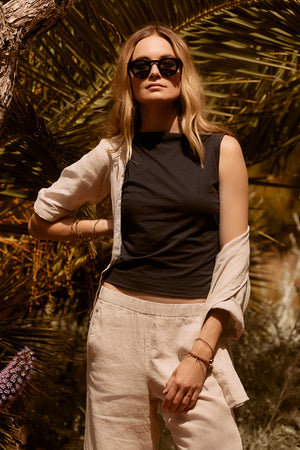 A woman in stylish sunglasses stands under a palm tree, wearing an Emilia Tank Top by Velvet by Graham & Spencer, a light open shirt, and beige pants.