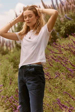 A young woman in a Velvet by Graham & Spencer Ryan Tee and blue jeans stands among purple flowers, with her arms raised adjusting her hair.