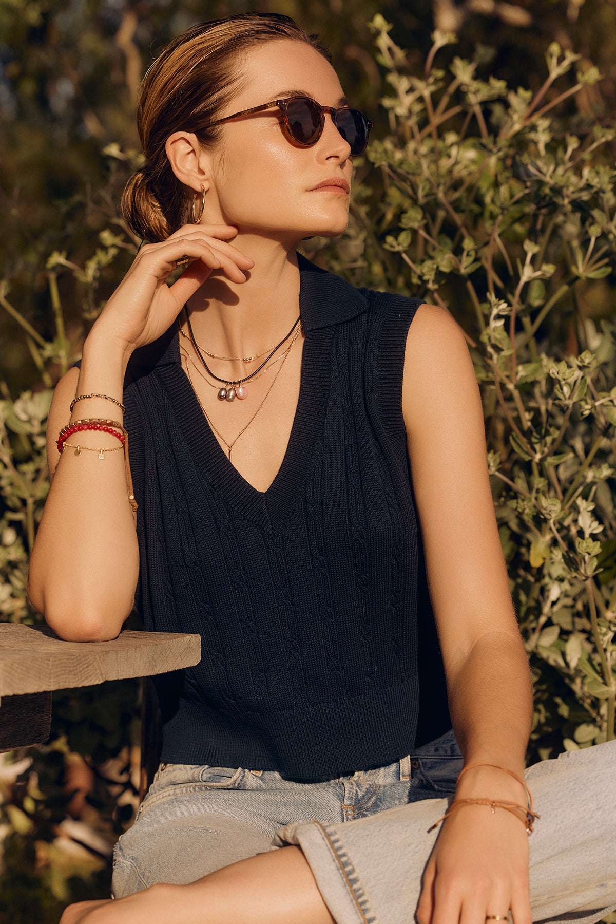 A woman wearing Velvet by Graham & Spencer sunglasses and a Wendy sweater vest sits outdoors, her hand on her neck, surrounded by green foliage.-36909792952513