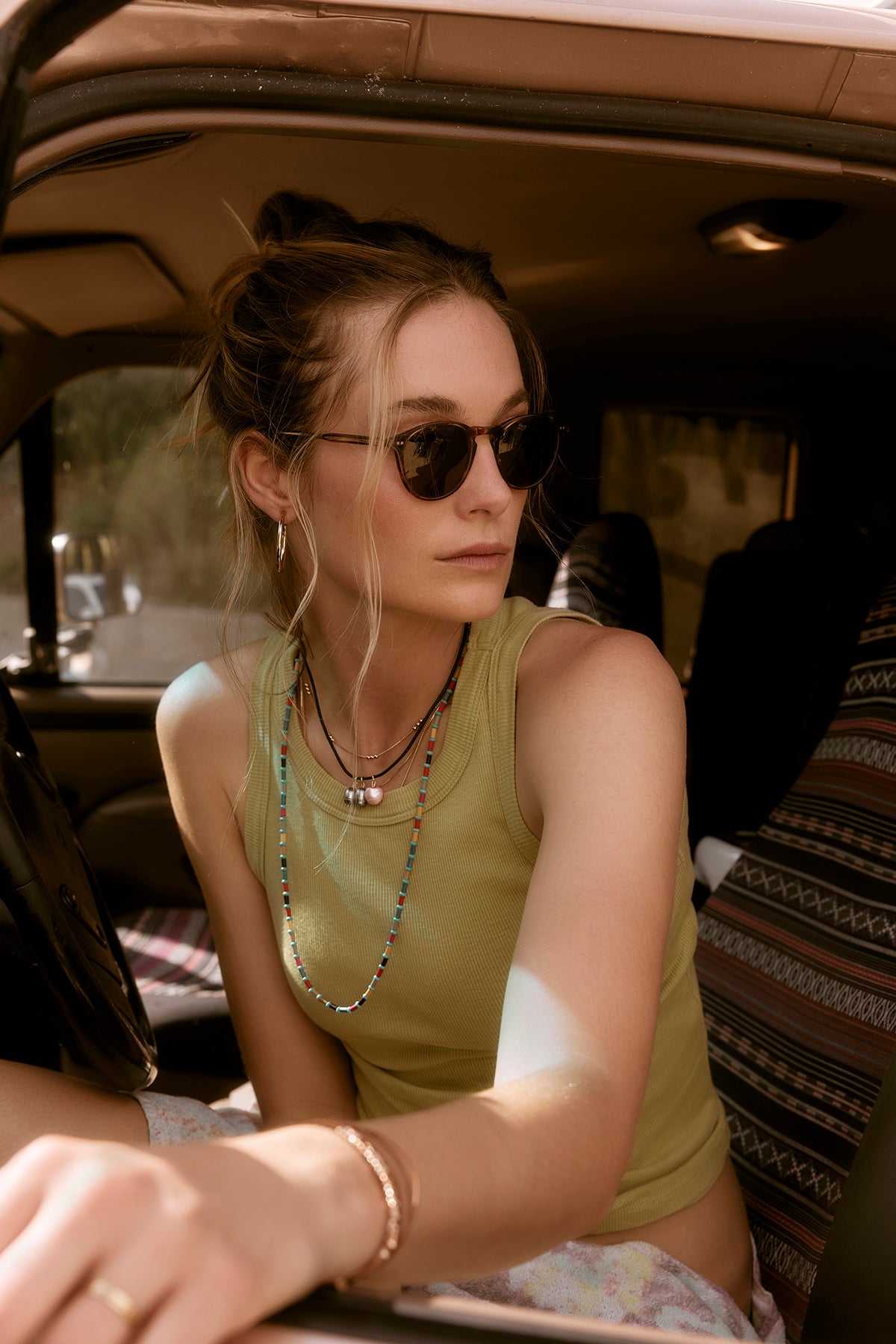   A young woman wearing sunglasses and a Velvet by Graham & Spencer NOLA tank top sitting inside a car, bathed in soft sunlight, looking to the side. 