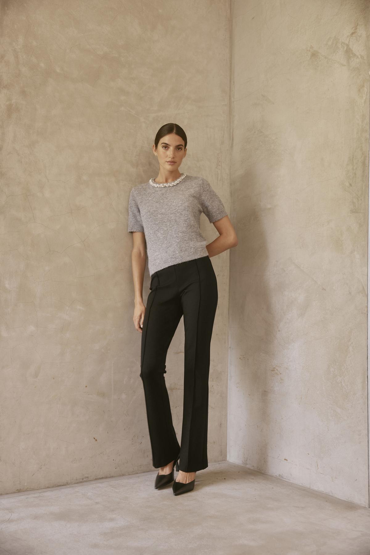   A person wearing the ALBEE PONTE PANT from Velvet by Graham & Spencer, characterized by its black color and elastic waist, stands leaning against a textured beige wall while sporting a gray top. 