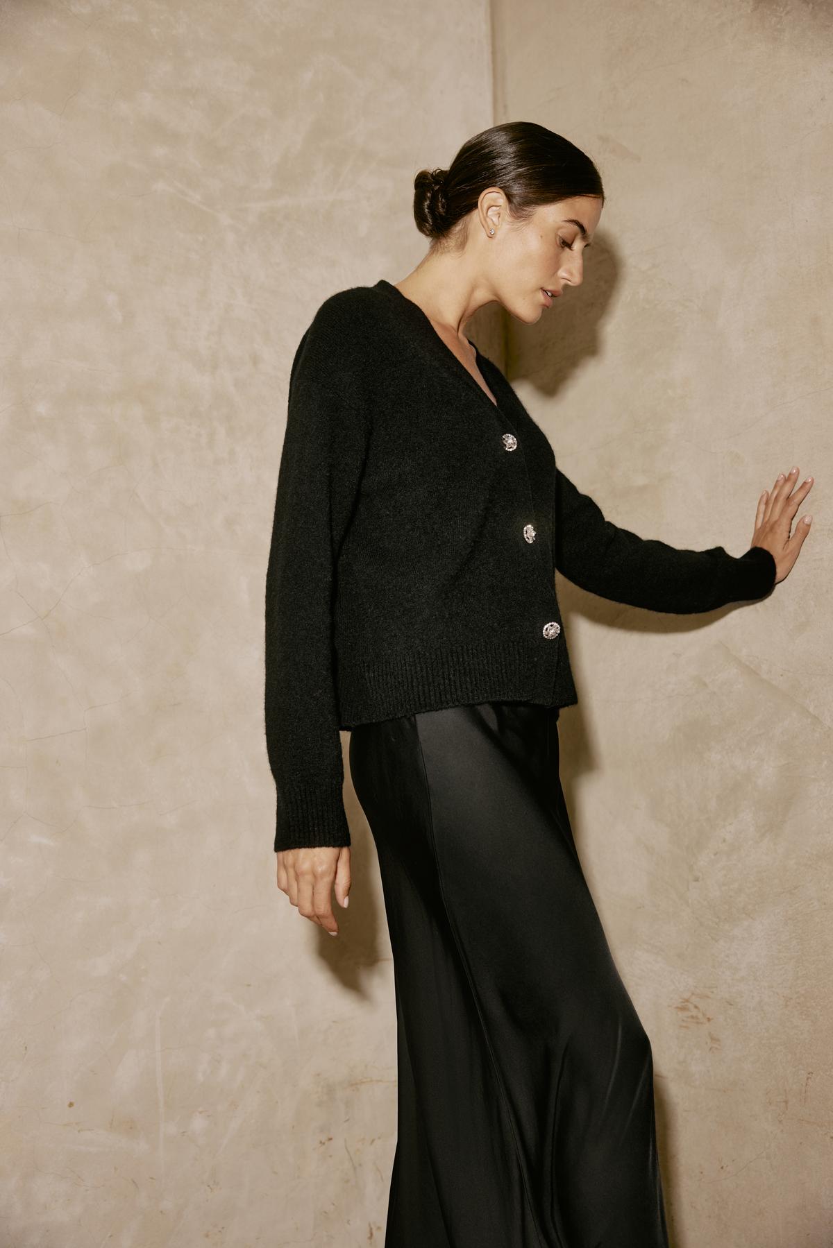   A person is standing against a beige wall, with one hand touching it, wearing a black sweater and the CADENCE SATIN SKIRT by Velvet by Graham & Spencer, featuring an elastic waist. 