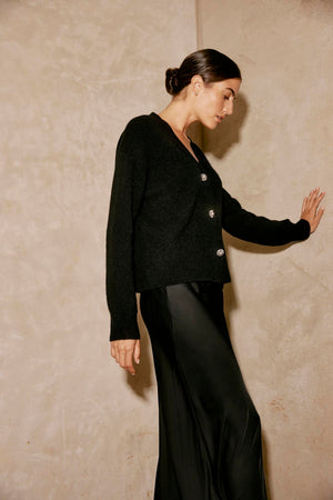 A person is standing against a beige wall, with one hand touching it, wearing a black sweater and the CADENCE SATIN SKIRT by Velvet by Graham & Spencer, featuring an elastic waist.