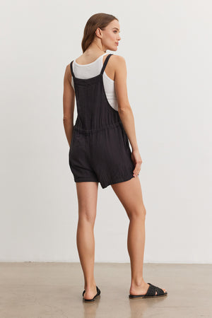 A woman stands in a studio, wearing a white tank top under black pinstriped Cyndee Cotton Gauze romper with a drawstring waist, paired with black sandals. She is facing away from the camera. (Brand: Velvet by Graham & Spencer)