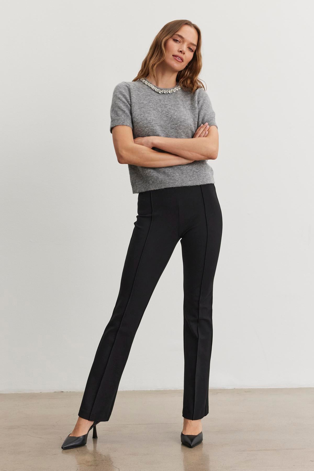   A person stands with arms crossed, wearing a grey short-sleeve top and the ALBEE PONTE PANT by Velvet by Graham & Spencer, featuring an elastic waist. They are in front of a plain, light-colored wall. 