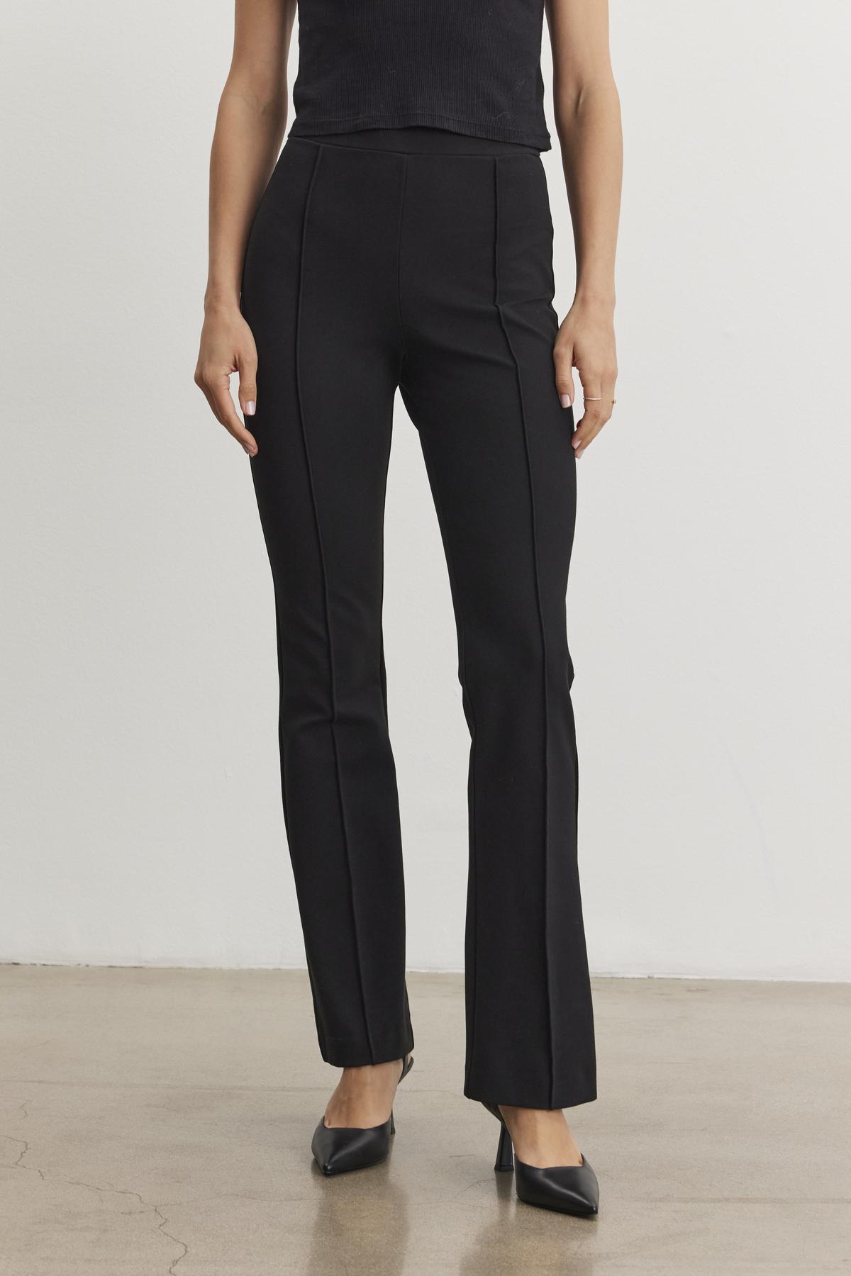 The individual is dressed in the ALBEE PONTE PANT from Velvet by Graham & Spencer, featuring a high-waisted and boot-cut silhouette, paired with a sleeveless top and black pointed shoes on a plain floor background.-38246314279105