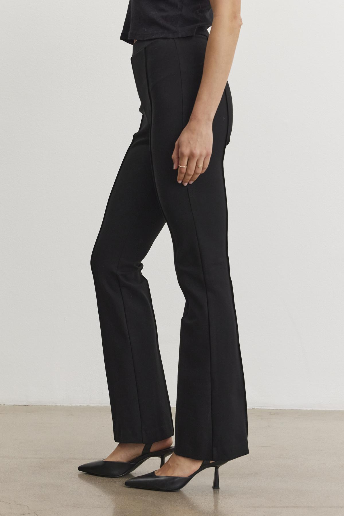   A person wearing the ALBEE PONTE PANT by Velvet by Graham & Spencer, featuring a stylish black bell-bottom design and an elastic waist, stands sideways on a plain floor in sleek black high heels. 