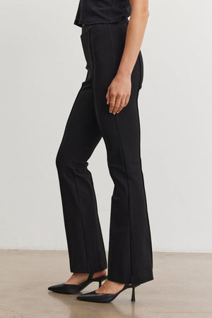 A person wearing the ALBEE PONTE PANT by Velvet by Graham & Spencer, featuring a stylish black bell-bottom design and an elastic waist, stands sideways on a plain floor in sleek black high heels.