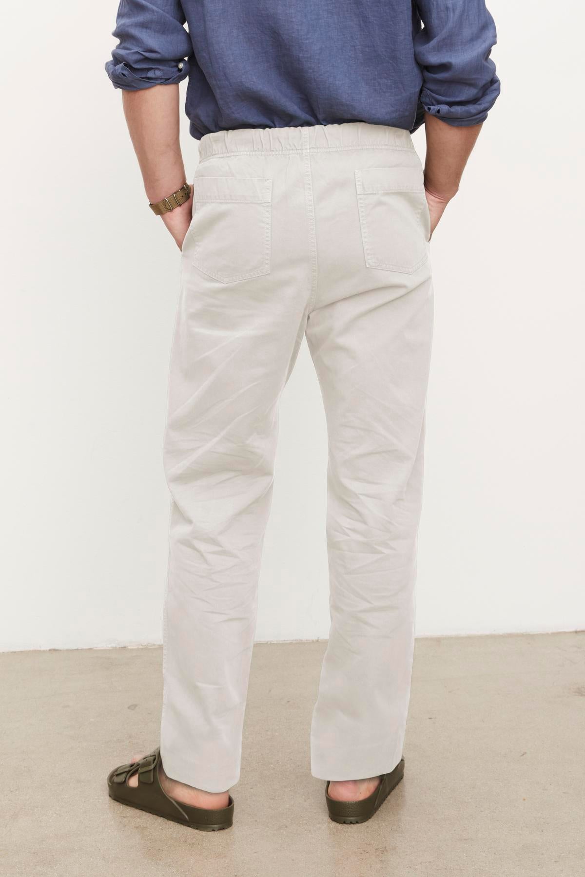   A person standing with their back to the camera, wearing light-colored cotton sanded twill Branson Pants by Velvet by Graham & Spencer and a blue shirt with rolled-up sleeves. 