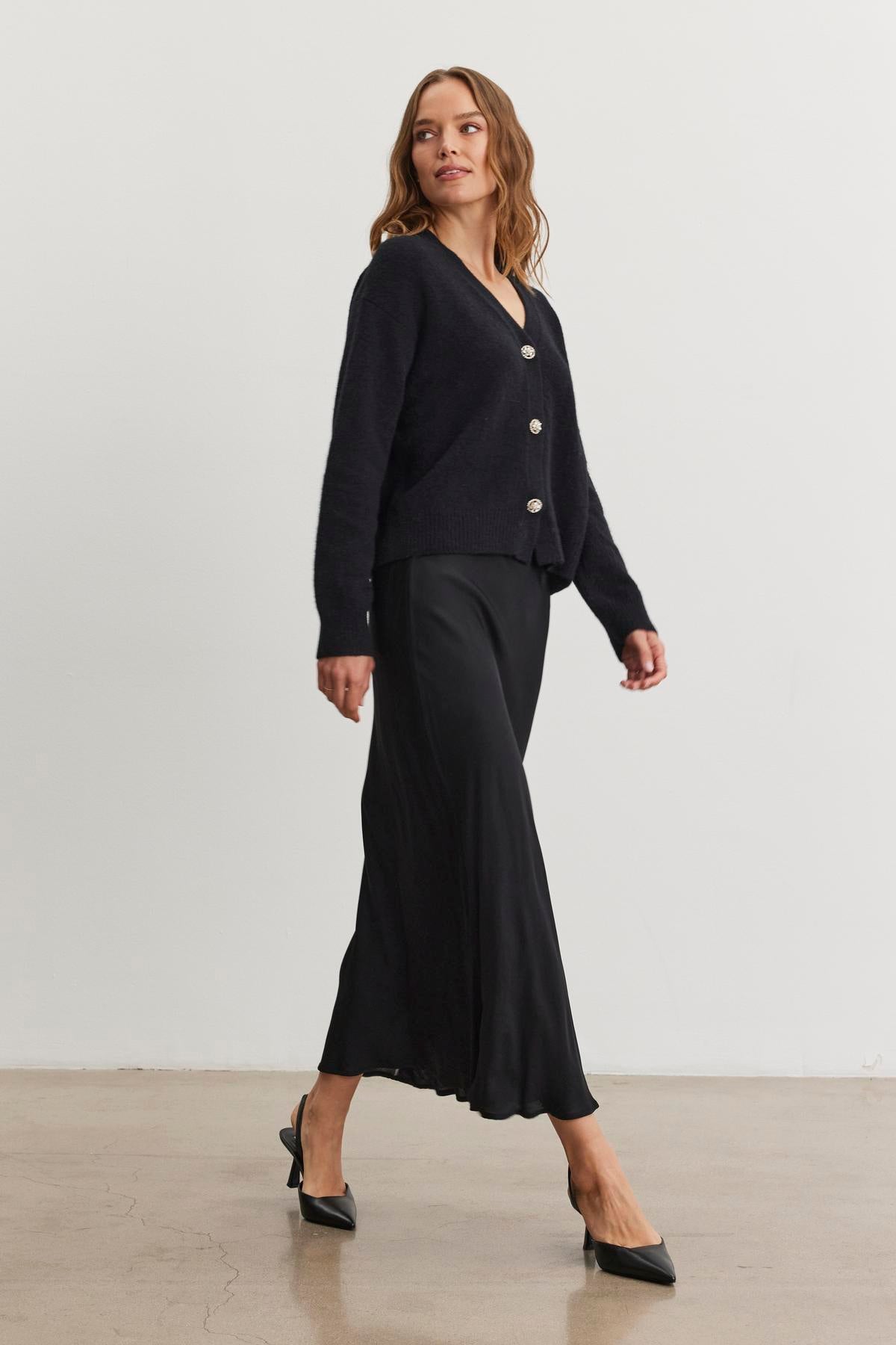   A person wearing a viscose cardigan and the CADENCE SATIN SKIRT from Velvet by Graham & Spencer, paired with black heels, walks on a light-colored floor. 