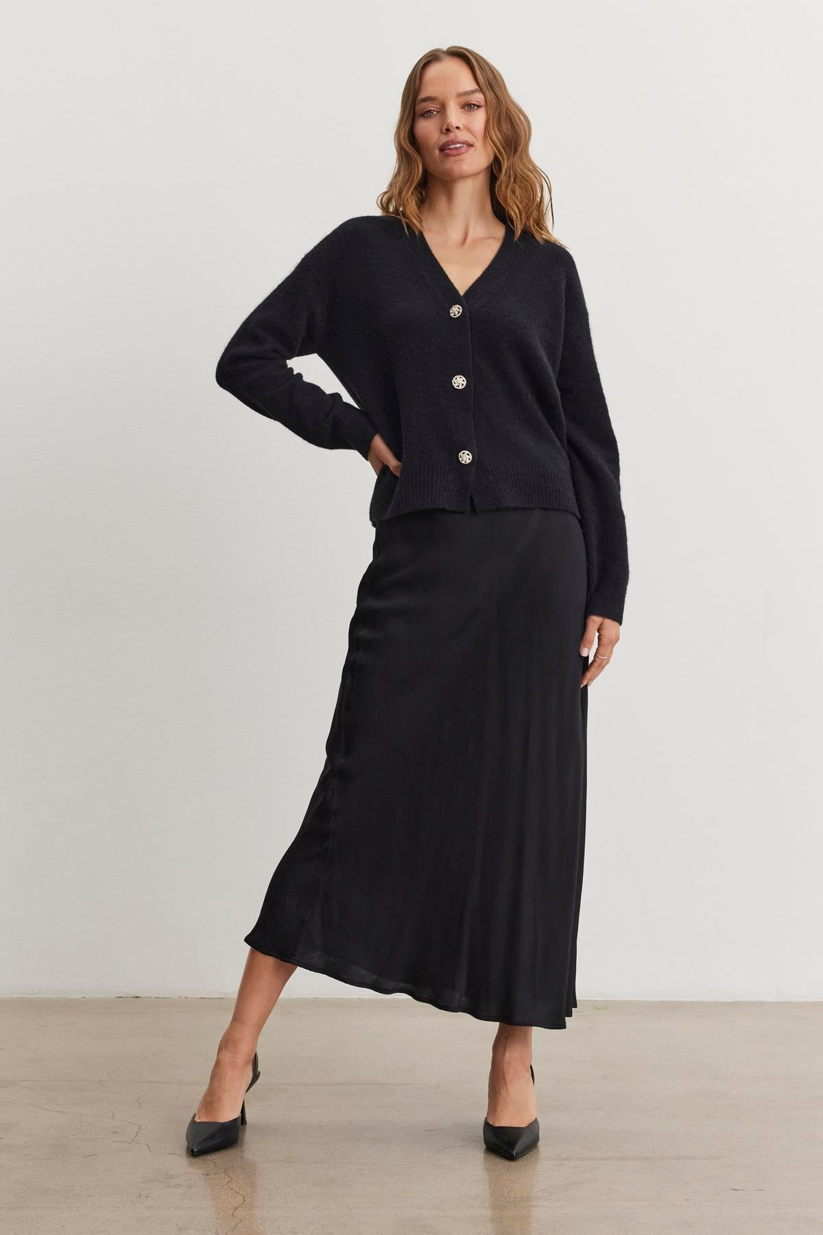 A woman stands confidently on a plain background, wearing the Cadence Satin Skirt by Velvet by Graham & Spencer, a long black skirt with an elastic waist that adds sophistication with its subtle sheen of satin viscose, as she poses with one hand on her hip.-38246369591489