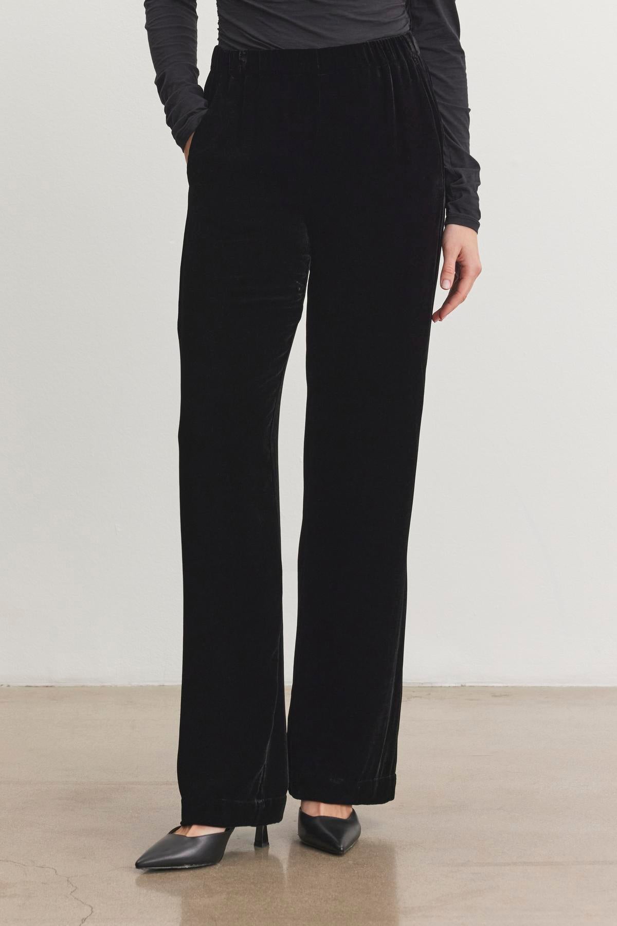   A person wearing Velvet by Graham & Spencer's SORINE SILK VELVET PANT, a long-sleeve top, and black heeled shoes, standing on a polished floor. 