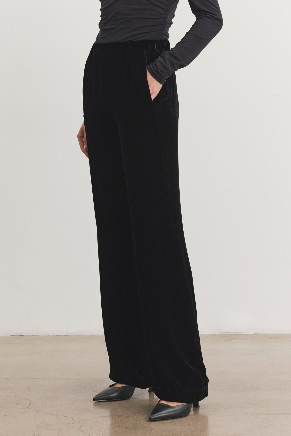   A person wears the SORINE Silk Velvet Pant by Velvet by Graham & Spencer, paired with a long-sleeve top and black pointed shoes, standing on a polished floor. 