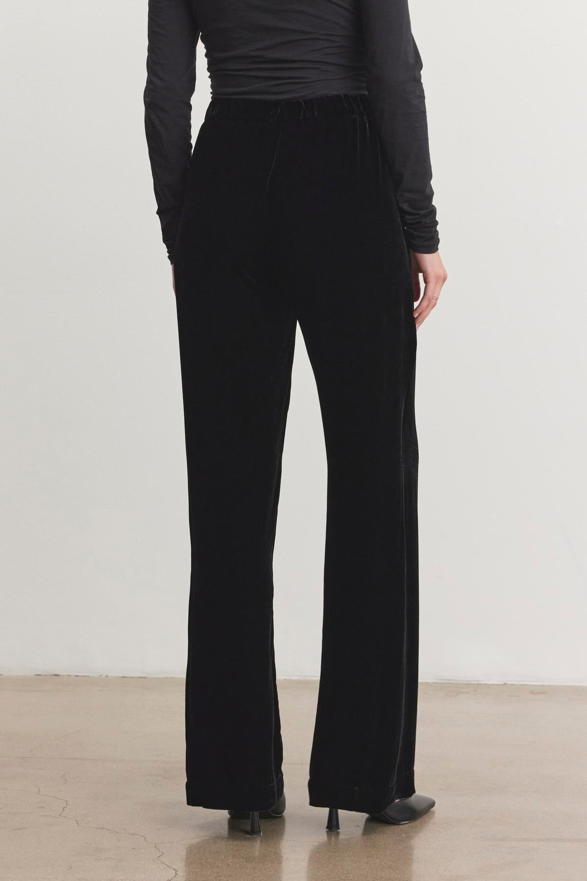   A person dressed in a long-sleeve black top and the SORINE SILK VELVET PANT by Velvet by Graham & Spencer, paired with heels, is standing on a polished floor. 