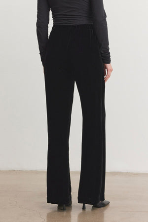 A person dressed in a long-sleeve black top and the SORINE SILK VELVET PANT by Velvet by Graham & Spencer, paired with heels, is standing on a polished floor.