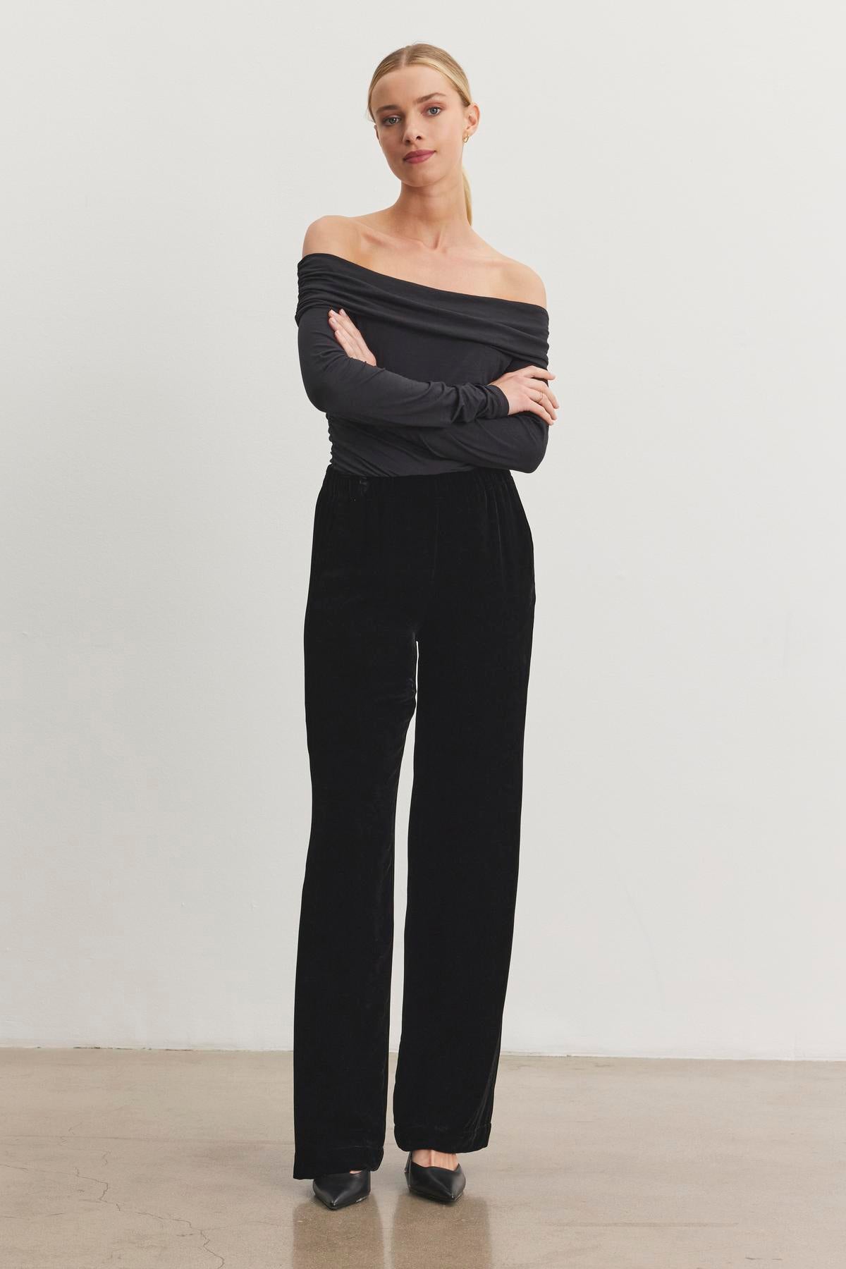   A person wearing an off-shoulder top and the SORINE SILK VELVET PANT by Velvet by Graham & Spencer stands on a plain background with arms crossed. 