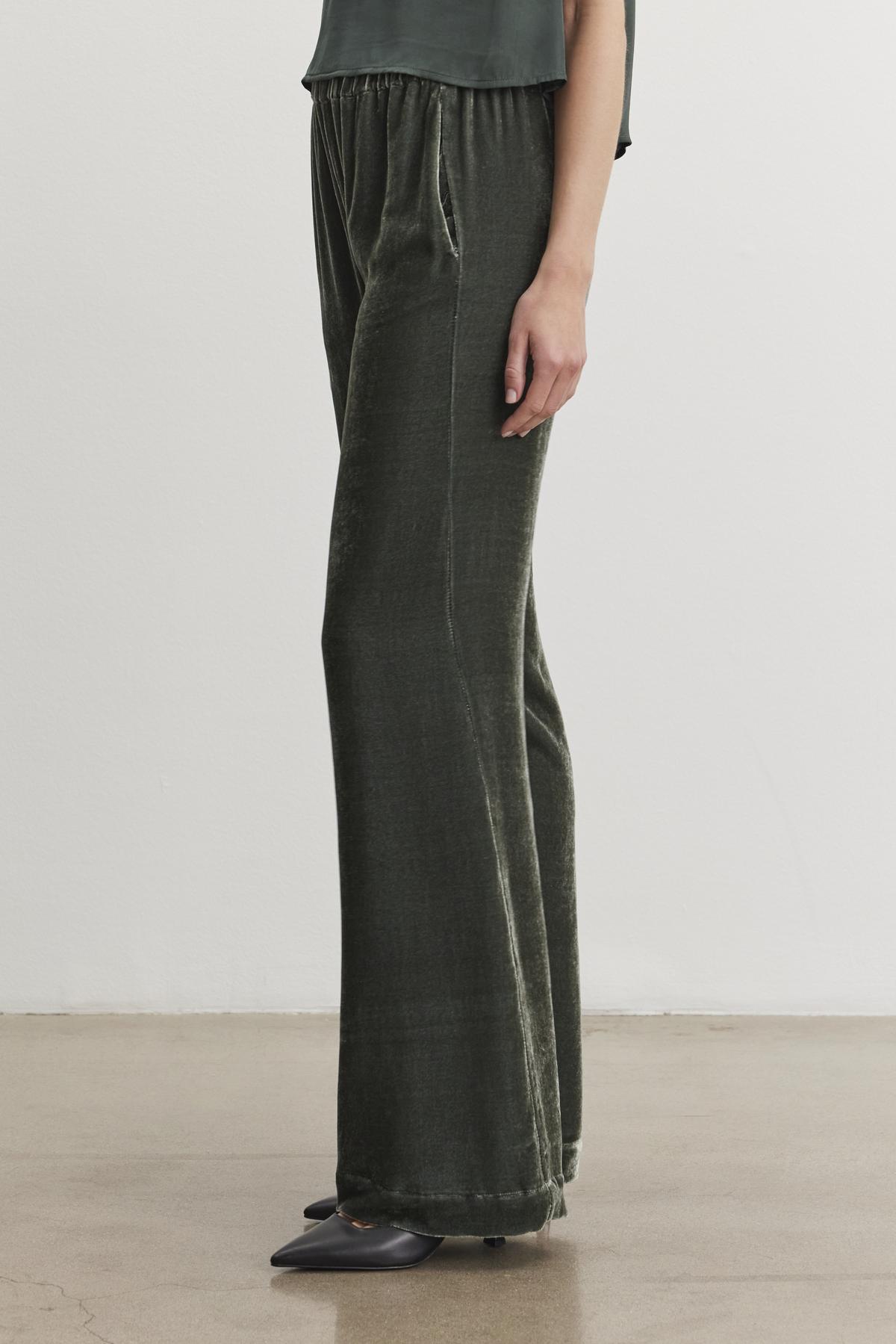   A person is standing indoors against a plain wall, wearing the SORINE SILK VELVET PANT by Velvet by Graham & Spencer paired with black pointed shoes. 