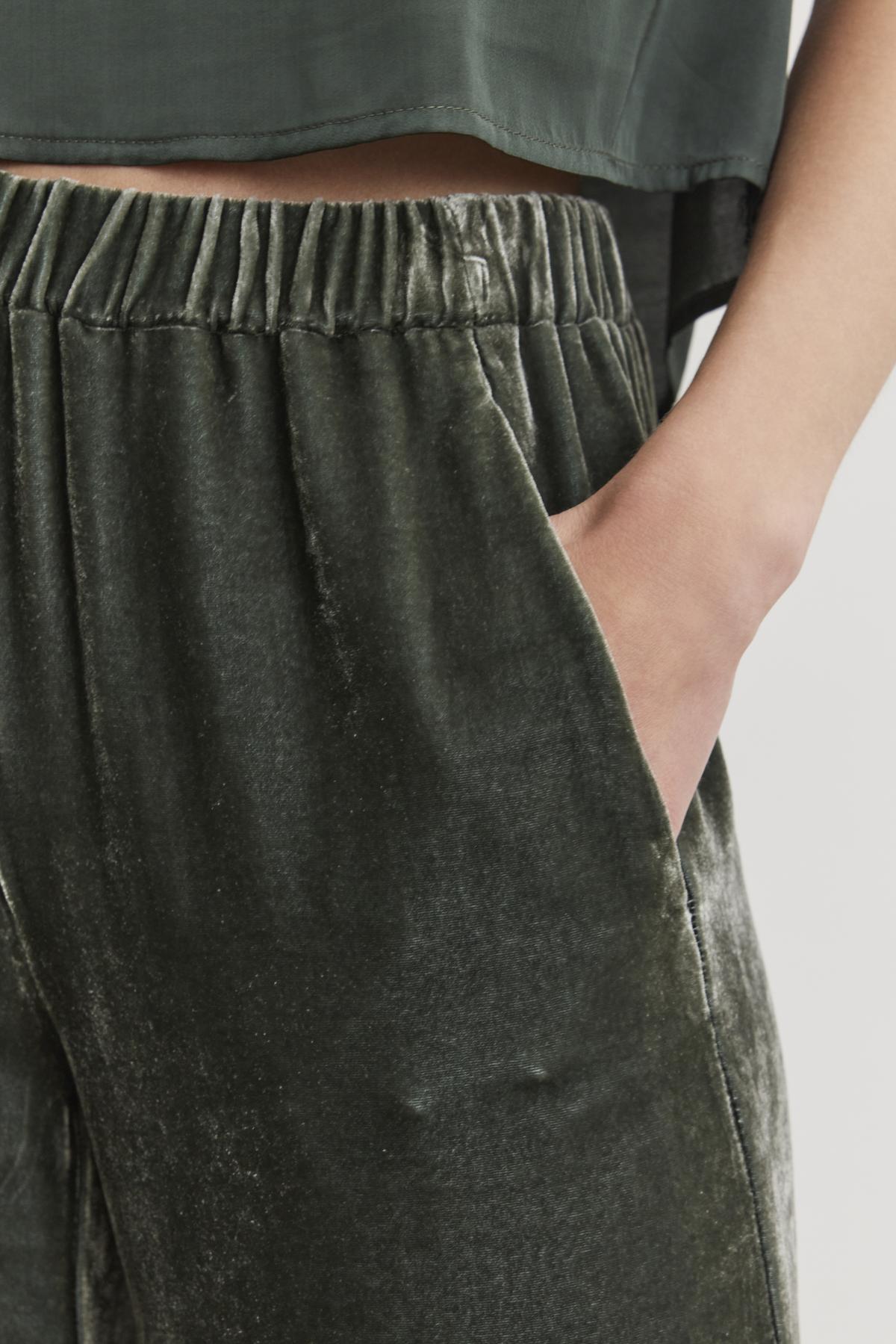   Close-up of a person wearing the SORINE SILK VELVET PANT by Velvet by Graham & Spencer in dark green, with a hand in the pocket, paired with a matching green top. 