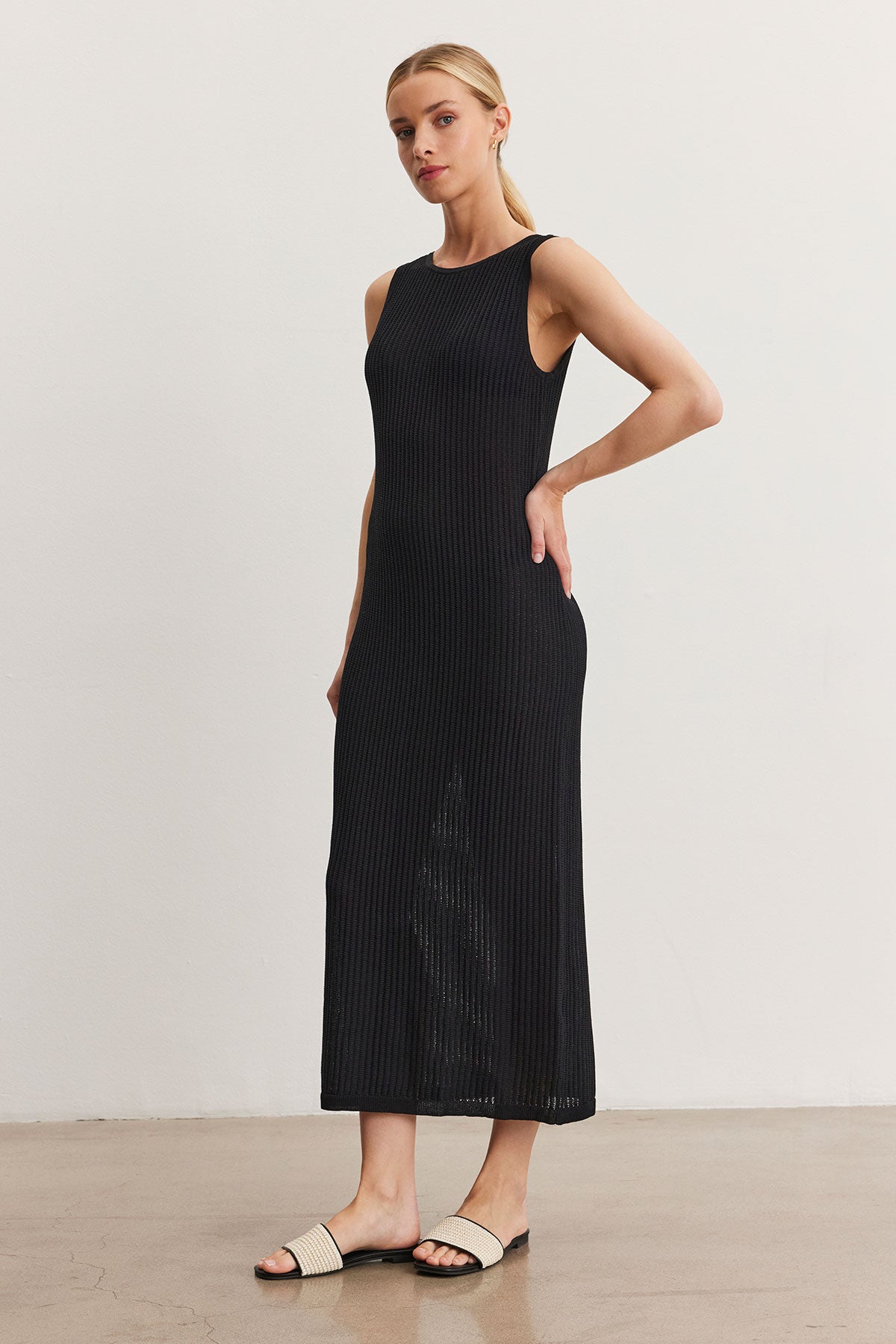   A woman in a Velvet by Graham & Spencer CALLEN DRESS and white sandals stands in a minimalistic room, posing with her hand on her hip. 