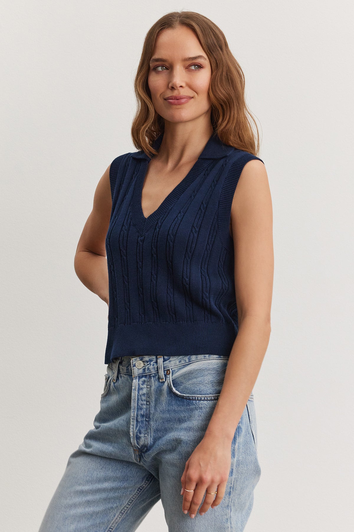   A woman in a Velvet by Graham & Spencer Wendy sweater vest and light blue jeans standing against a plain background, smiling gently. 