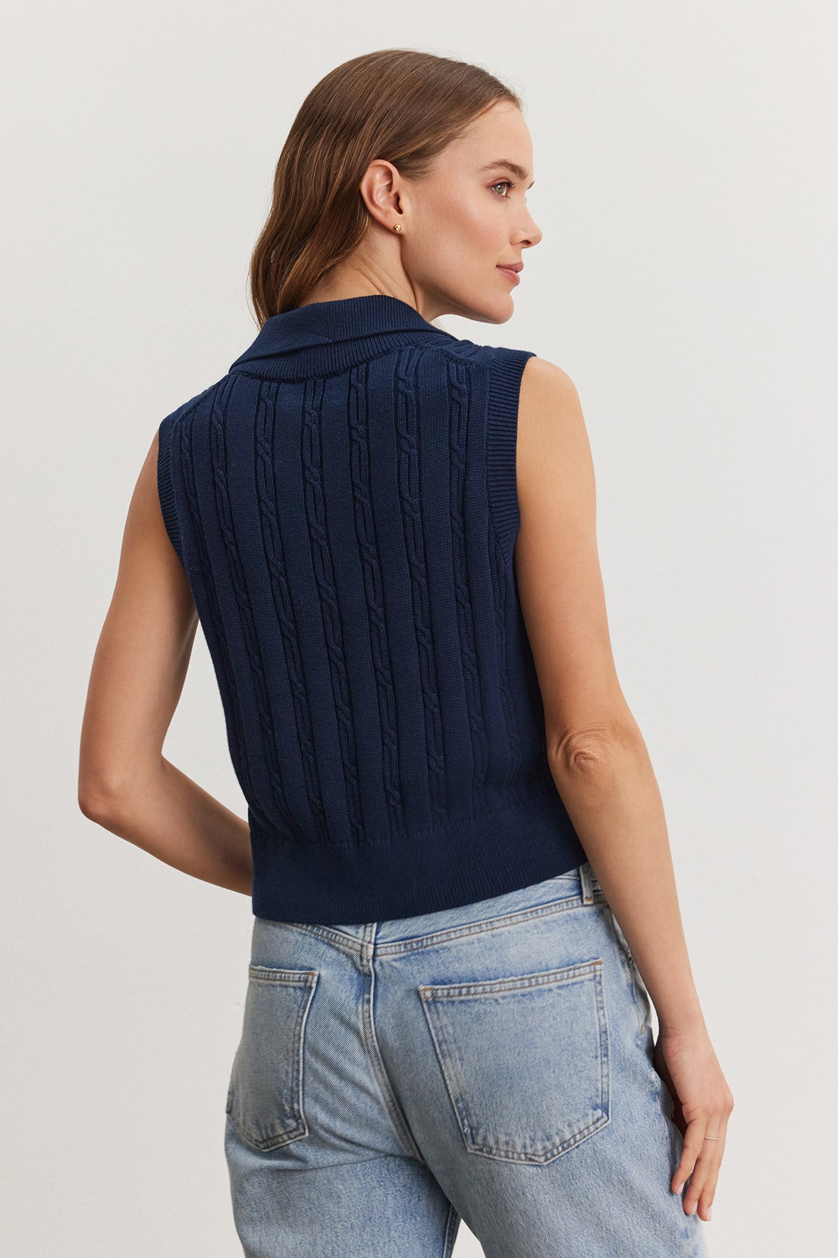   Woman standing facing away from the camera, wearing a blue Velvet by Graham & Spencer Wendy Sweater Vest and light blue jeans, focusing on the sweater's cable knit design. 