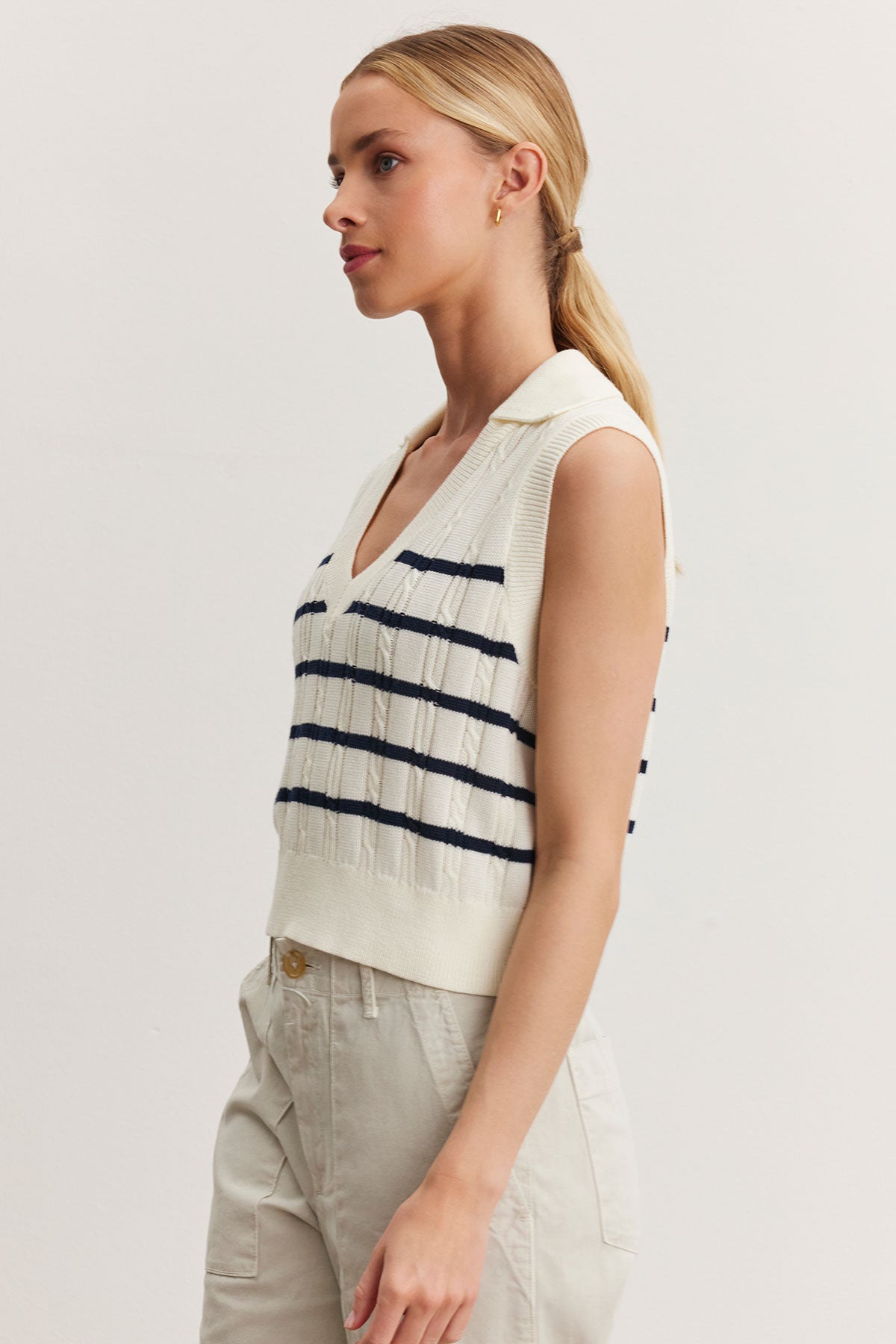   A woman poses in a Velvet by Graham & Spencer Wendy sweater vest with blue stripes, paired with light beige trousers, against a white backdrop. 