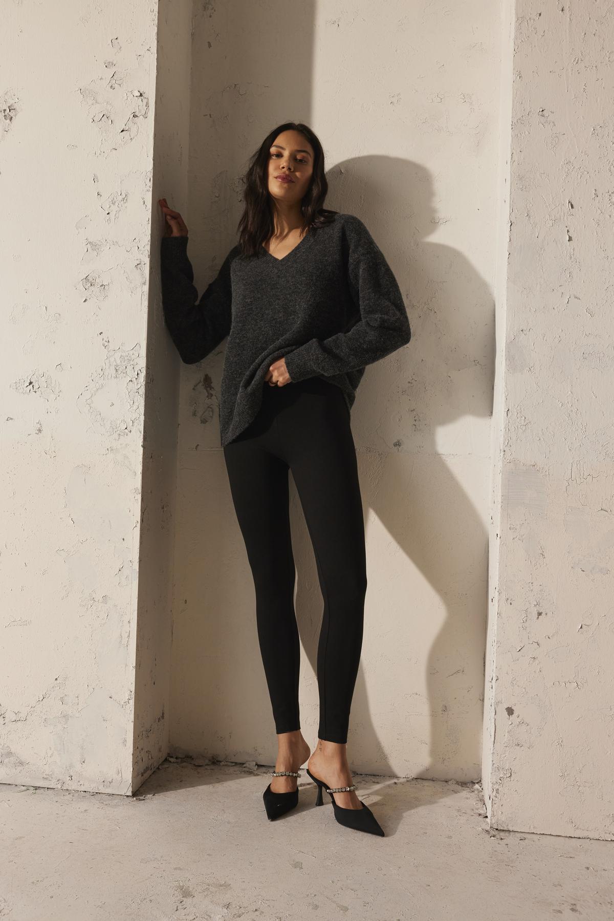   A person wearing the KELSA ALPACA SWEATER by Velvet by Graham & Spencer, along with black leggings and pointed black shoes, is leaning against a textured white wall. 