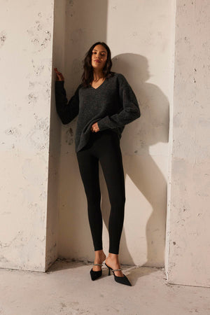 A person wearing the KELSA ALPACA SWEATER by Velvet by Graham & Spencer, along with black leggings and pointed black shoes, is leaning against a textured white wall.