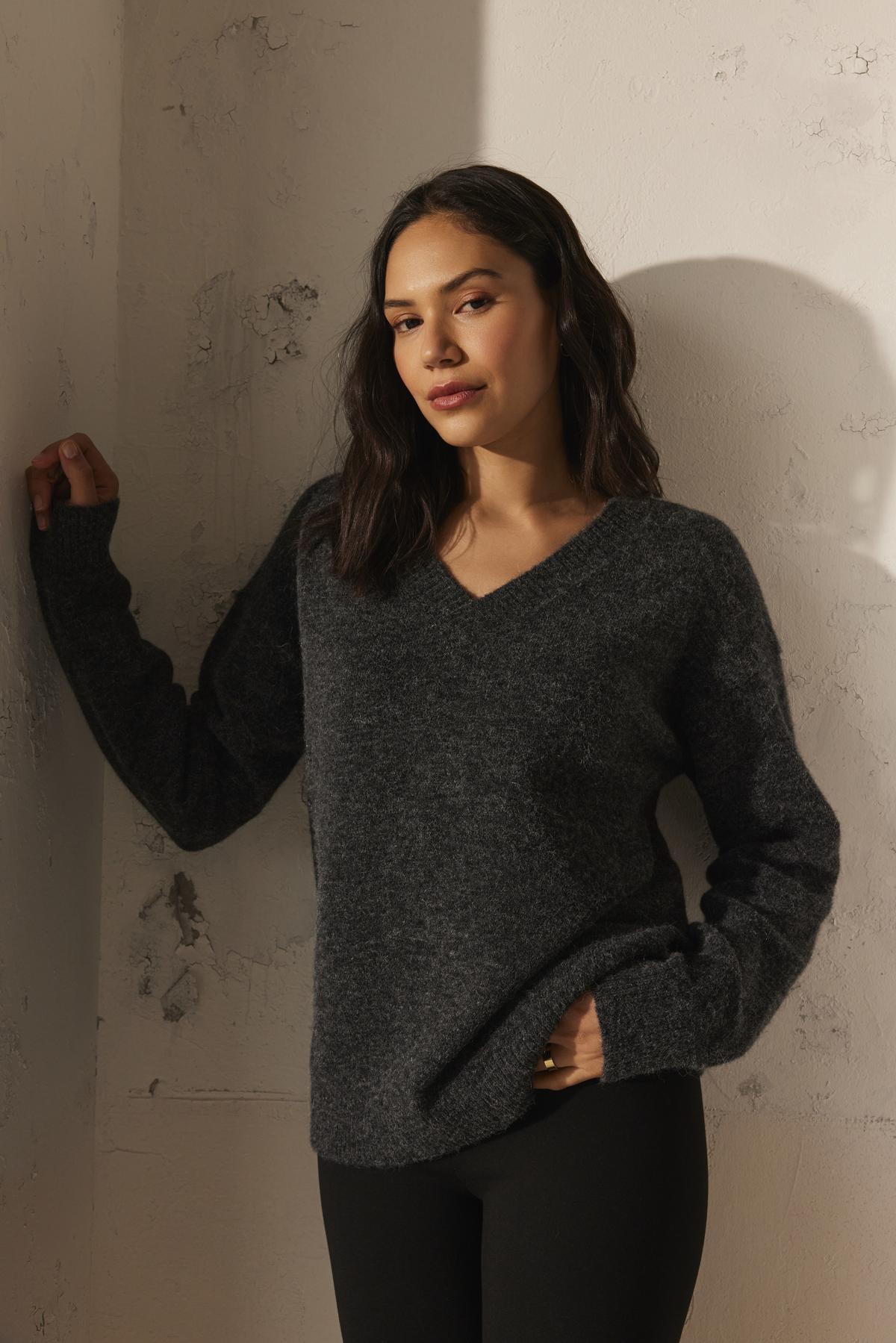   A woman wearing the KELSA ALPACA SWEATER by Velvet by Graham & Spencer and black pants leans against a textured wall, looking directly at the camera. 