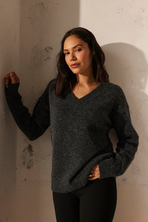 A woman wearing the KELSA ALPACA SWEATER by Velvet by Graham & Spencer and black pants leans against a textured wall, looking directly at the camera.